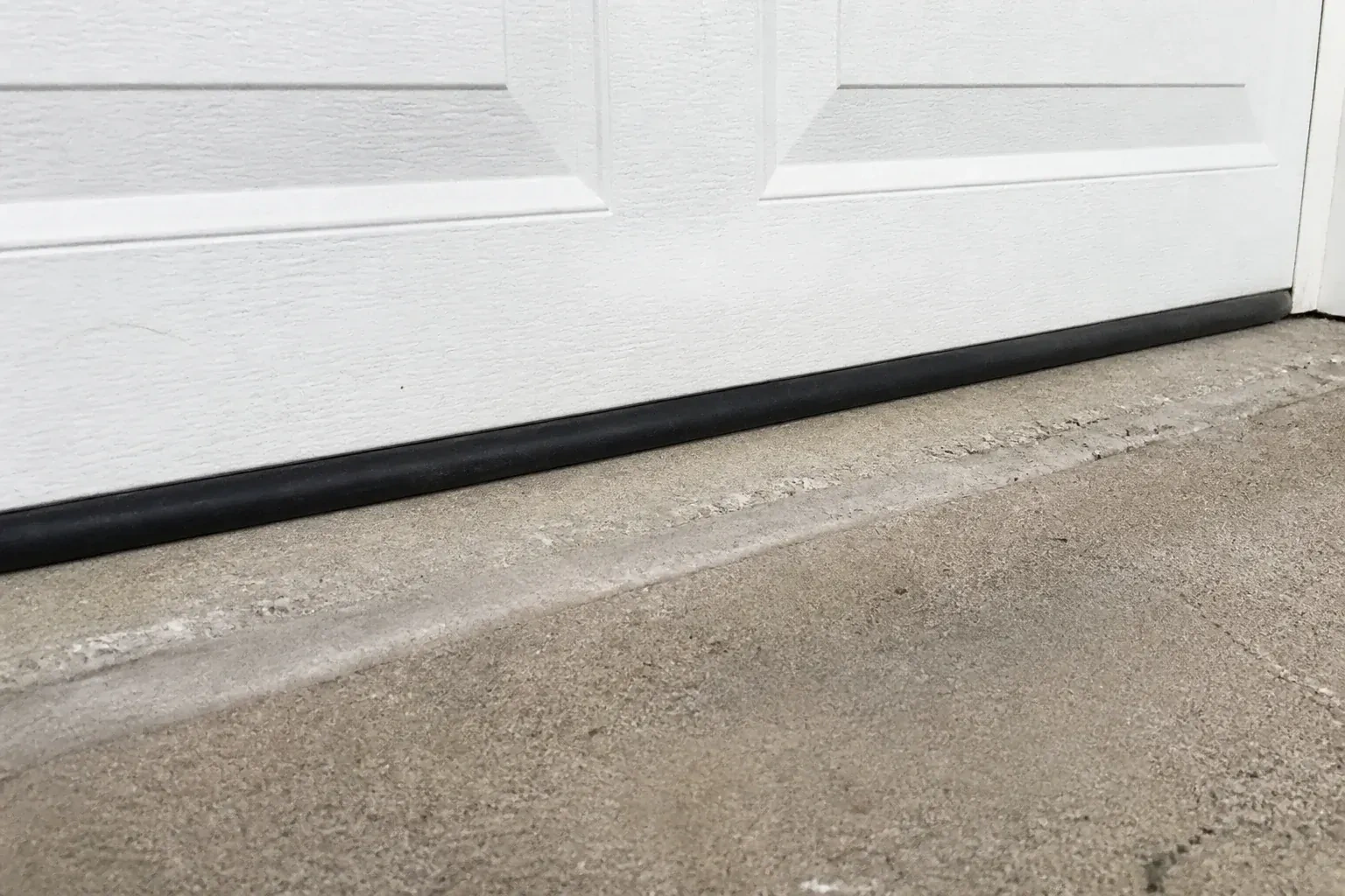 Black bottom seal along a closed white garage door at a concrete threshold. — Garage Door Bottom Seal Replacement, Brampton