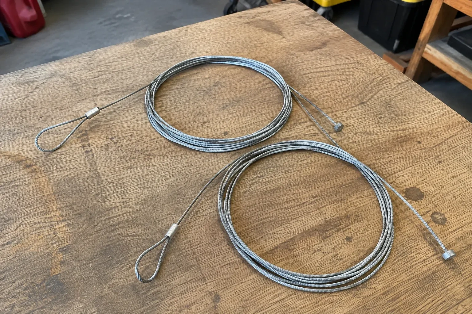 Two coiled steel cables with loop and stop ends on a workbench. — Garage Door Cable Replacement, Brampton